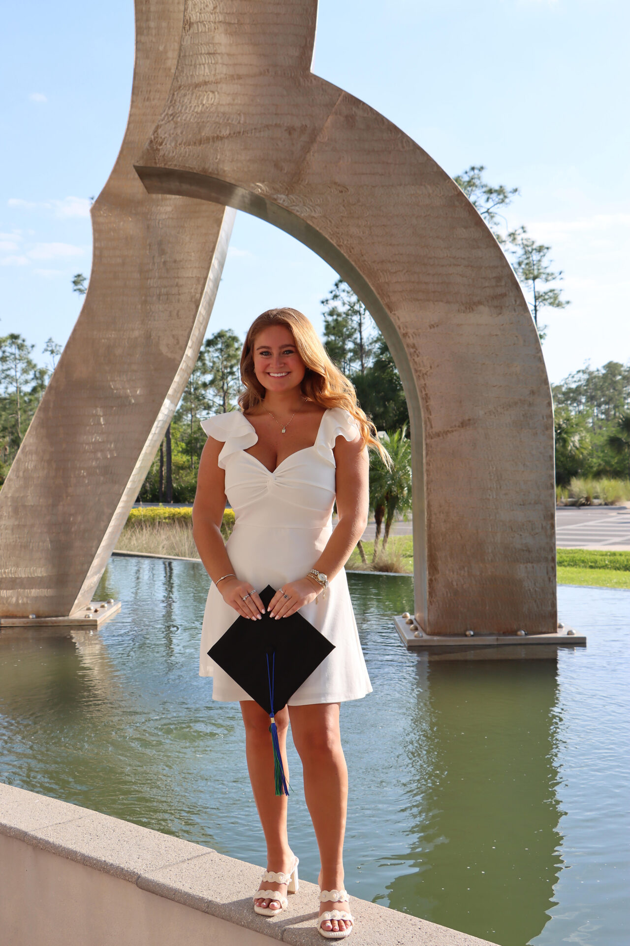 Smiling young woman in a white dress holding a graduation cap near a large arch-shaped sculpture by a reflecting pool.