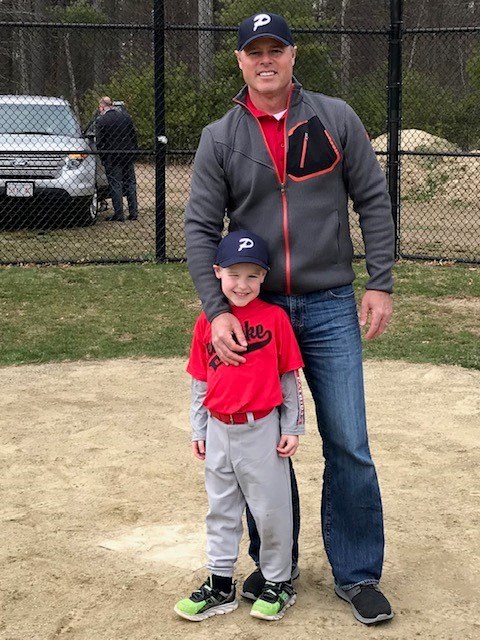 Father and son pose for a photo on a baseball field; the man wears a gray jacket and cap, with his arm around a smiling young player in a red jersey and gray pants, chain-link fence in the background.